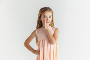 Stylish little smiling girl posing in dress isolated on white studio background. Caucasian blonde female model. Human emotions, facial expression, childhood. Whispering a secret, smiling.