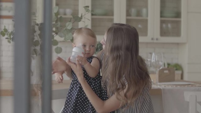 Beautiful smiling young woman showing her baby girl how to drink from the baby bottle in the kitchen. Concept of a happy family, one child, motherhood, love. Slow motion