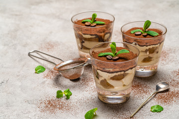 Classic tiramisu dessert in a glass cup and strainer with cocoa powder on concrete background