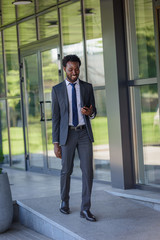 smiling african american businessman holding smartphone while walking along office buiding