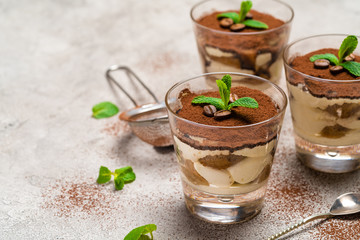 Classic tiramisu dessert in a glass cup and strainer with cocoa powder on concrete background