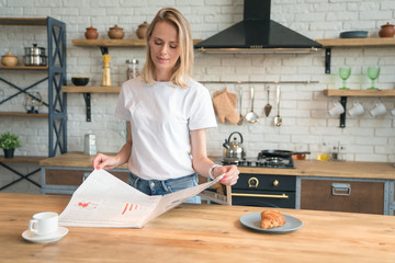 young beautiful woman is reading her morning press while having breakfast in the kitchen. coffee with croissants. wearing white shirt and jeans