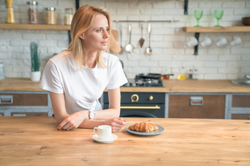 young beautiful woman is reading her morning press while having breakfast in the kitchen. coffee with croissants. wearing white shirt and jeans