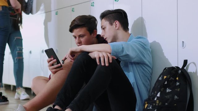 Two friendly classmates spend time together use a smartphone while sitting by lockers in school corridor during a break.