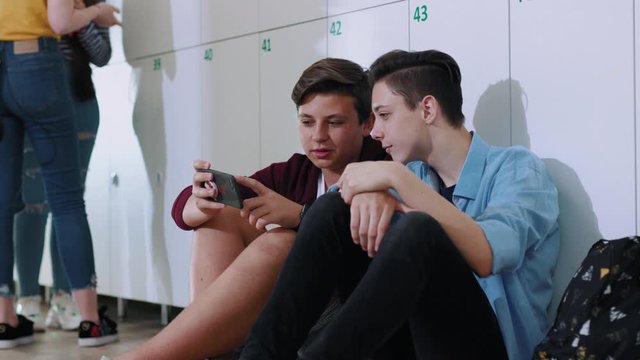 Modern teen students watching funny videos on smartphone talking together sitting by lockers in a school corridor. Fun school break. Friendship concept.
