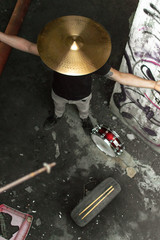 Drummer in grunge place with a cymbal of a drum in his head, snare drum and drumstick in the floor. Rock drummer in alternative urban place.