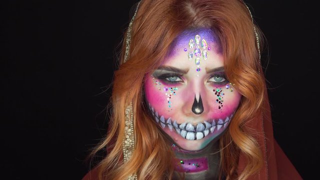 Gothic style, mystical make-up in image of pink sugar sweet skull, girl ready for carnival in honor of Day of Dead, lady with rhinestones and sparkles on face in dark room smiles with mockery