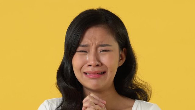Disappointed Asian woman crying with tears on her face in front of camera against yellow background for sad emotion and breakdown concepts