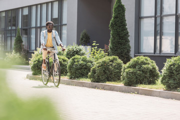 selective focus of stylish african american man riding bicycle along street with green bushes