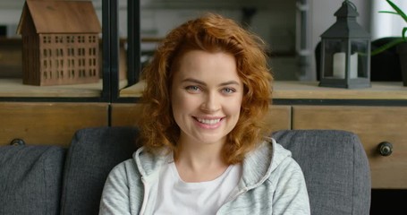 Portrait of young woman changing mood from sad to happy and positive, sitting on sofa at home, smiling, looking at camera, full joy, red curly hair, blue eyes, Caucasian. 4K, shot on RED camera.