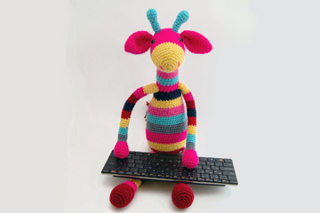 A multi-colored doll Amigurumi giraffe sits at a personal computer with a keyboard.