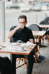 Stylish young brunette man sits in a cafe on the terrace and drinks coffee. Italian street