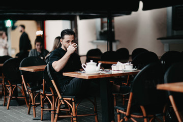 Stylish young brunette man sits in a cafe on the terrace and drinks coffee. Italian street