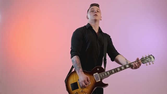 Handsome man in black shirt playing electric guitar with red and violet lights at background. Young rock musician with tattoo playing solo guitar. Medium shot