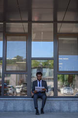 handsome african american businessman reading newspaper while sitting near office building with glass facade