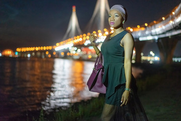 Fototapeta premium African fashion model by the water. Girl in a black dress. Black girl in the night city. African American on the waterfront. A woman looks into the distance.Girl is waiting for the return of boyfriend
