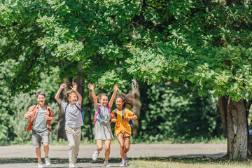 Obraz premium four happy multicultural schoolkids gesturing with raised hands while running in park