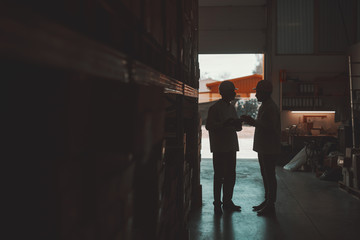 Silhouette of two hardworking warehouse workers standing in warehouse and checking inventory. Older one holding tablet while younger one looking at it.