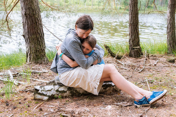 Naklejka na ściany i meble Mature middle-aged woman hugging her daughter in the forest to her breast - sitting on a tree trunk in the forest on the shore of the lake