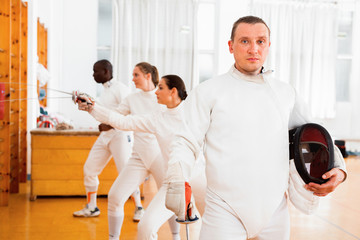 Active young male fencer in uniform standing with mask and foil at fencing room