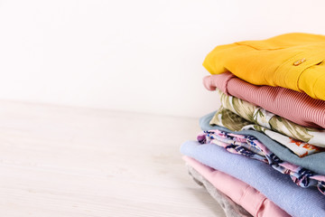 Stack of clean freshly laundered, neatly folded women's clothes on wooden table. Pile of shirts, dresses and sweaters on white board, concrete wall background. Copy space, close up, top view.