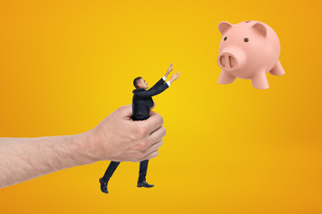 Big hand holding small businessman reaching out with his both hands for cute pink piggy bank floating in air on amber background.