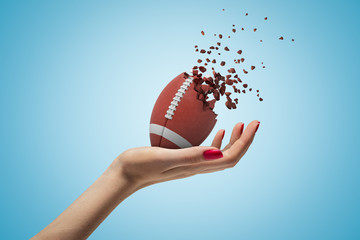 Female hand holding american football ball shattering into pieces on blue background