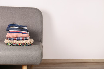 Bunch of clothing, knitted warm pastel color sweaters, different shirts and pants folded in stack on grey textile couch. Fall winter season apparel stacked on sofa. Close up, copy space, background.