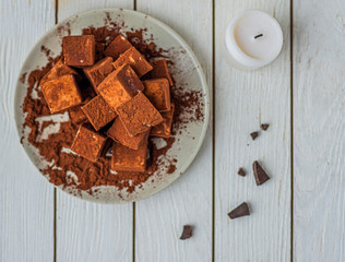 handmade souffle cubes covered with cocoa powder lie on a plate on a light wooden table, white candle and pieces of chocolate