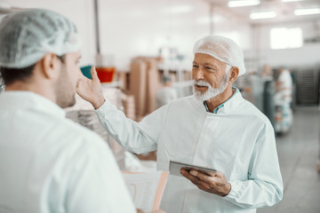 Two supervisors discussing about quality of food in food plant. Younger one holding folder with data while older one holding tablet. Both are dressed in white sterile uniforms and having hairnet.