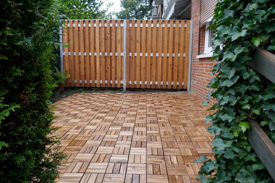Wooden partition and wooden tiles, two white garden chairs and a round table on the, with ivy and Thuja plants covered, terrace of a residential building