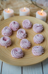 Merengue Marshmallow Zephyr lined in a circle on a bamboo plate on a white wooden background and three white burning candles. Top view. Pink sweet homemade zephyr or marshmallow.
