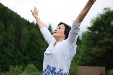 Senior woman doing a stretching exercise for the upper arms outside over landscape of forest and...