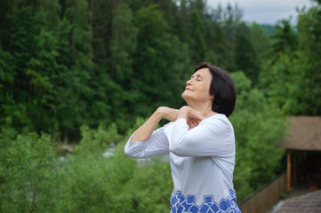 Senior woman doing a stretching exercise for the upper arms outside over landscape of forest and mountains