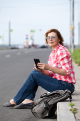 Smiling mature woman sitting on sloping curb, smartphone in her hands, looking far away, empty road
