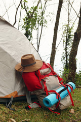 A backpack with a mat and a hat near to a tent on a grass in a camping on a background of forest.