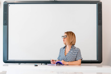 Young pensive attractive professor with glasses in formal clothes sitting near blackboard