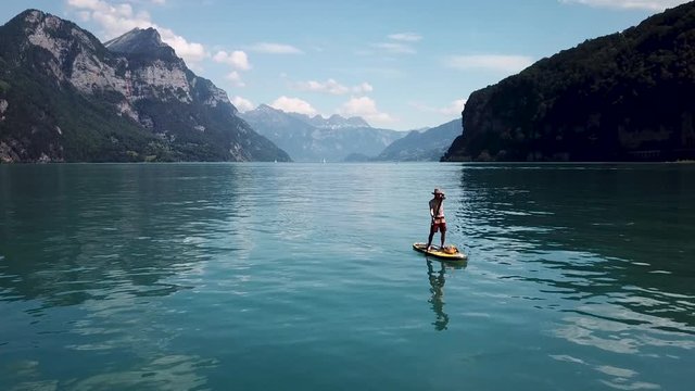 A white male is holding up his paddle on a stand up paddle, sup, the drone flies up to show him in the middle of a lake in Switzerland.
