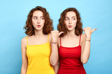 charming lovely puzzled two women demonstrating copyspace near shoulder isolated on blue background. twins showing aside. look here. gossip concept . body language