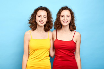 joyful identical positive stylish twins in red and yellow dresses looking at the camera. close up photo. isolated blue background, lifestyle, double beauty, people