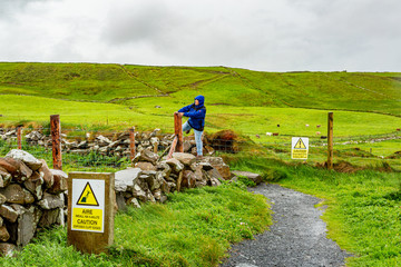 Happy woman jumping a fence on a rainy day in the coastal walk route from Doolin to the Cliffs of Moher, geosites and geopark, Wild Atlantic Way, spring day in county Clare in Ireland