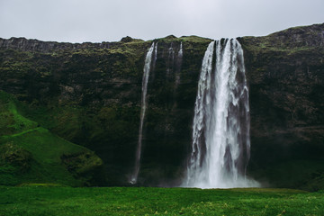 Famous Seljalandsfoss waterfall in Iceland. Saturated green grass, pale gray rocks, and heavy rainy sky