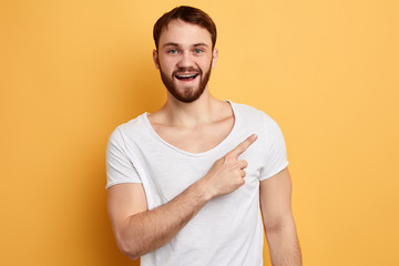 happy handsome young man in white t-shirt pointing finger away at copy space isolated over yellow background. close up photo. direction concept. this way