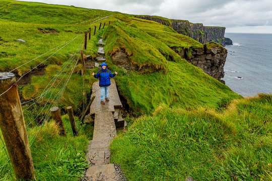 Irish landscape with cliffs and sea in background, female hiker on a bridge and stone coastal hiking trail from Doolin to Cliffs of Moher, rainy day in County Clare, Ireland. Wild Atlantic Way
