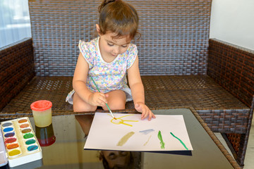Little child drawing a picture with paint and brush. Cute little brunette girl sitting among comfortable modern design garden furniture with couch, armchairs, and table arranged on a home patio.