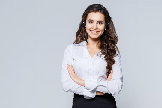 Young friendly operator woman agent with headsets standing near gray background. Call Center Service. Photo of customer support or sales agent in smart casual wear with crossed arms.