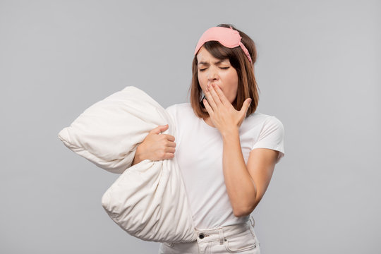 Young woman in sleepwear and mask covering her mouth while yawning