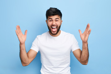 Cheerful handsome happy guy with wide opened mouth raising his hands up, having eyes full of happiness, rejoicing his achievements.