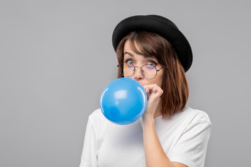 Young brunette female in white t-shirt and black hat blowing blue balloon