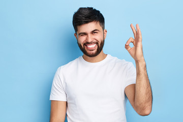 happy funny bearded man winking his eye and showing ok sign, isolated on blue background,...